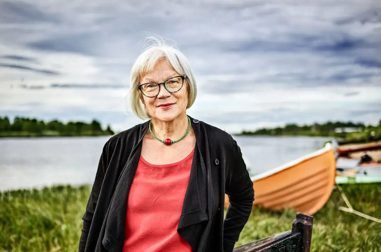 Britta Marakatt Labba, med sitt vita h&aring;r och glas&ouml;gon, st&aring;r vid en sj&ouml; med b&aring;tar i bakgrunden.