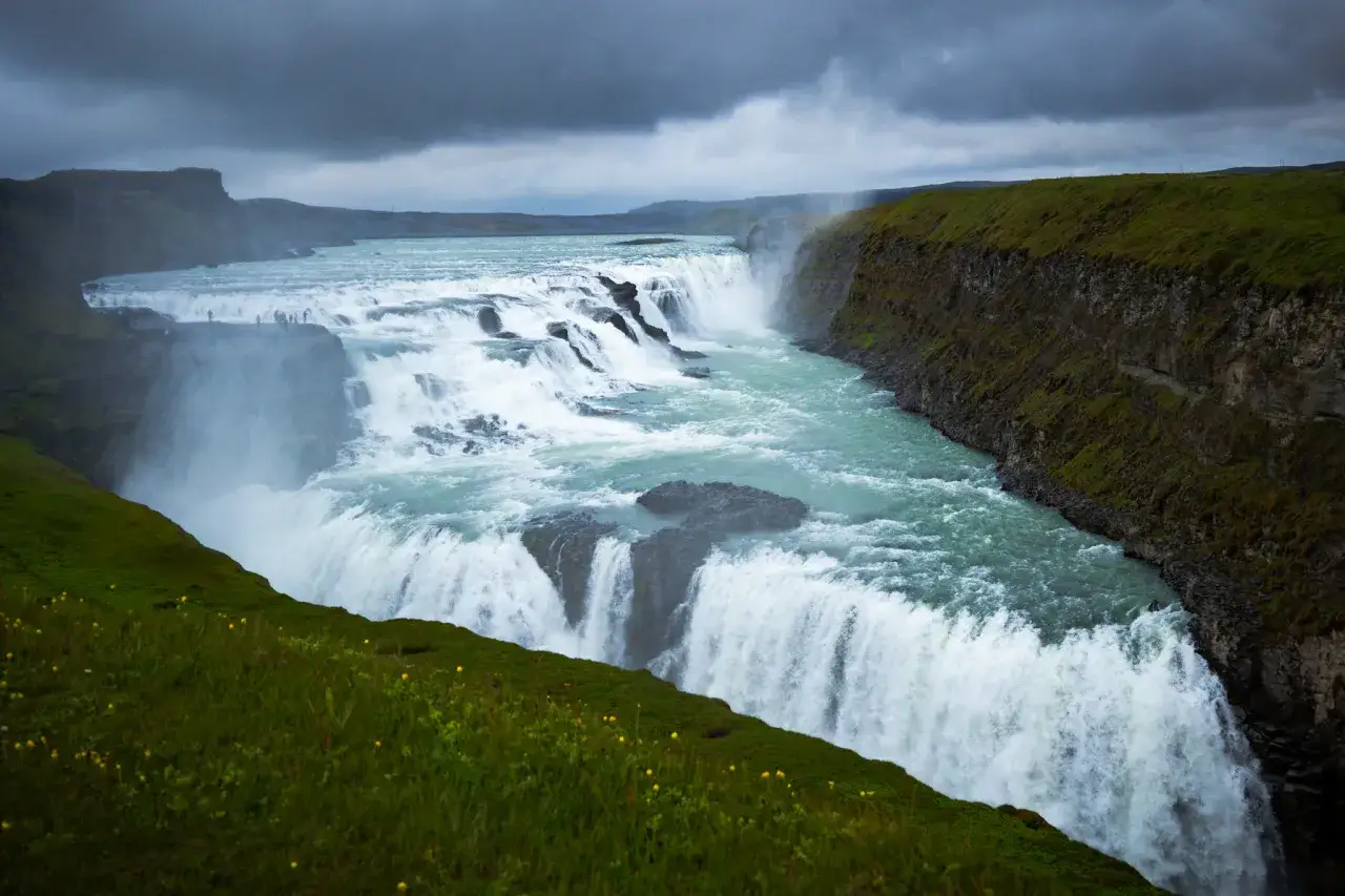 Potężny wodospad Gullfoss, punkt kulminacyjny tygodniowej wycieczki po Islandii, w otoczeniu zielonych wzgórz i dramatycznego, pochmurnego nieba.