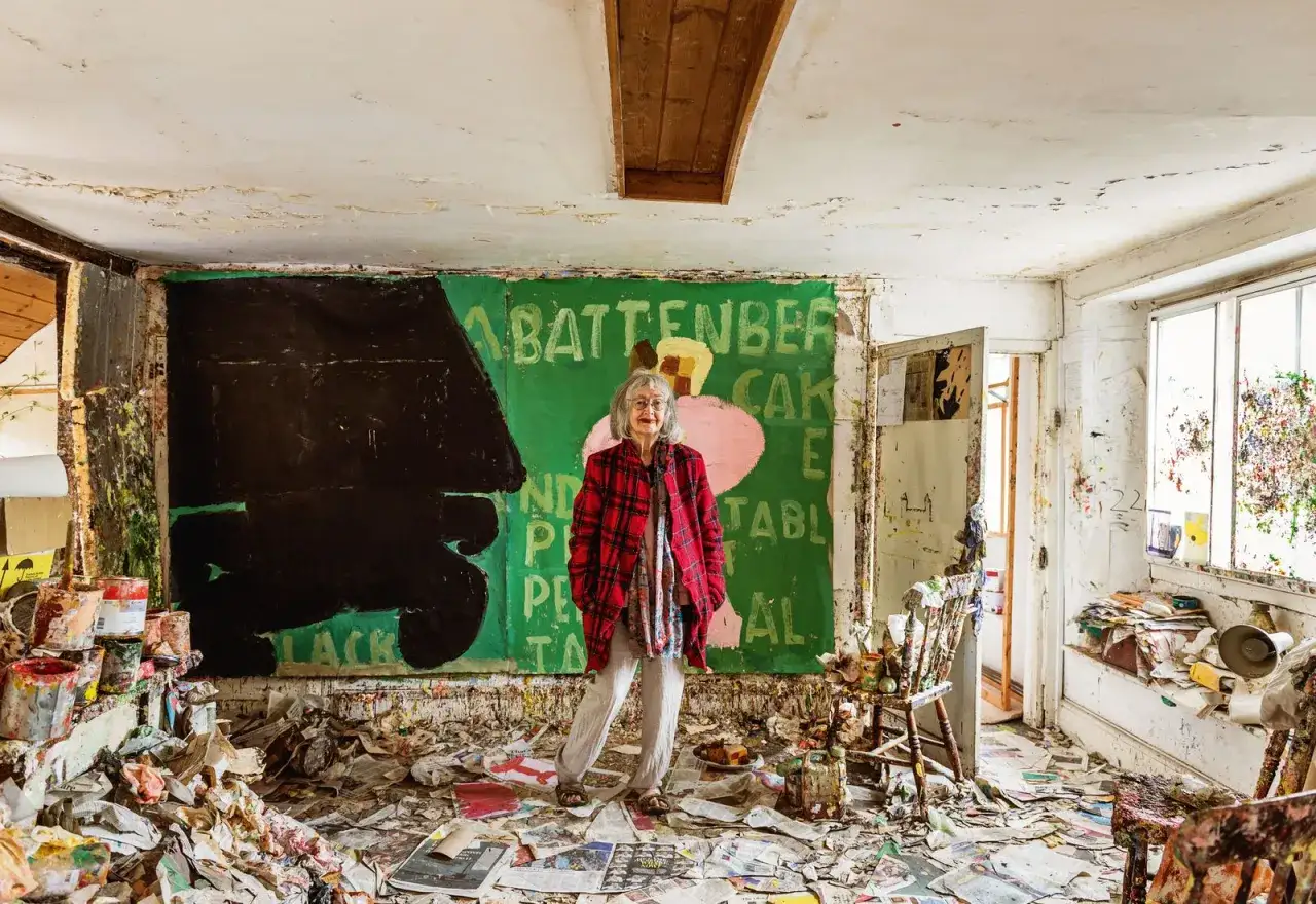 Rose Wylie stands in her paint-splattered studio, a vibrant green canvas behind her depicting a Battenberg cake.