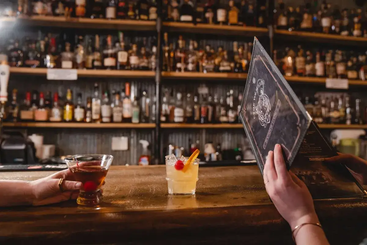 Due mani sul bancone di un bar, una tiene un cocktail con bourbon, l'altra sfoglia il menu.