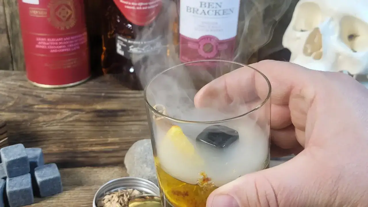 Dym unoszący się nad drinkami z whisky, z pomarańczą i kamieniem w szkle. W tle butelki i czaszka.