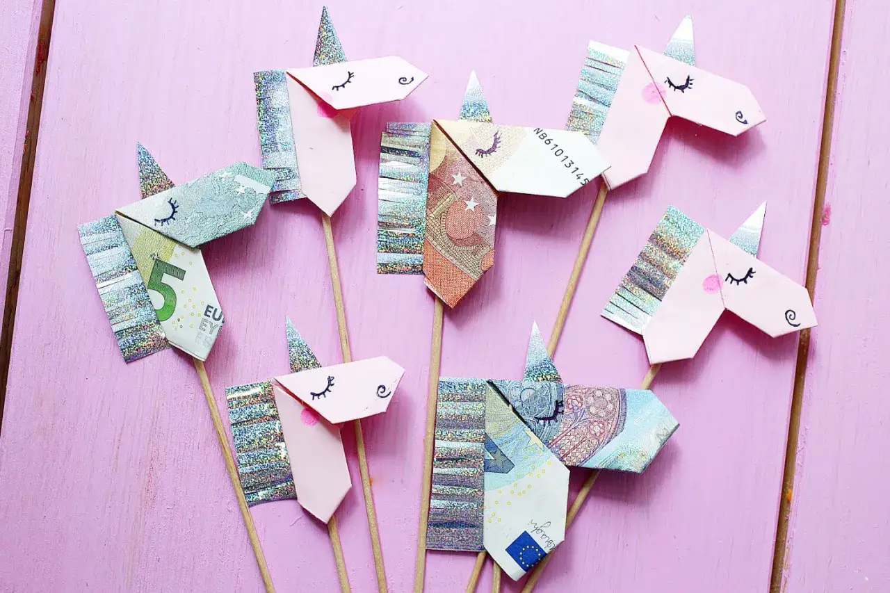 Origami-Einhörner aus gefalteten Euro-Scheinen auf rosa Holzuntergrund. Ein kreatives Geld falten für besondere Anlässe.