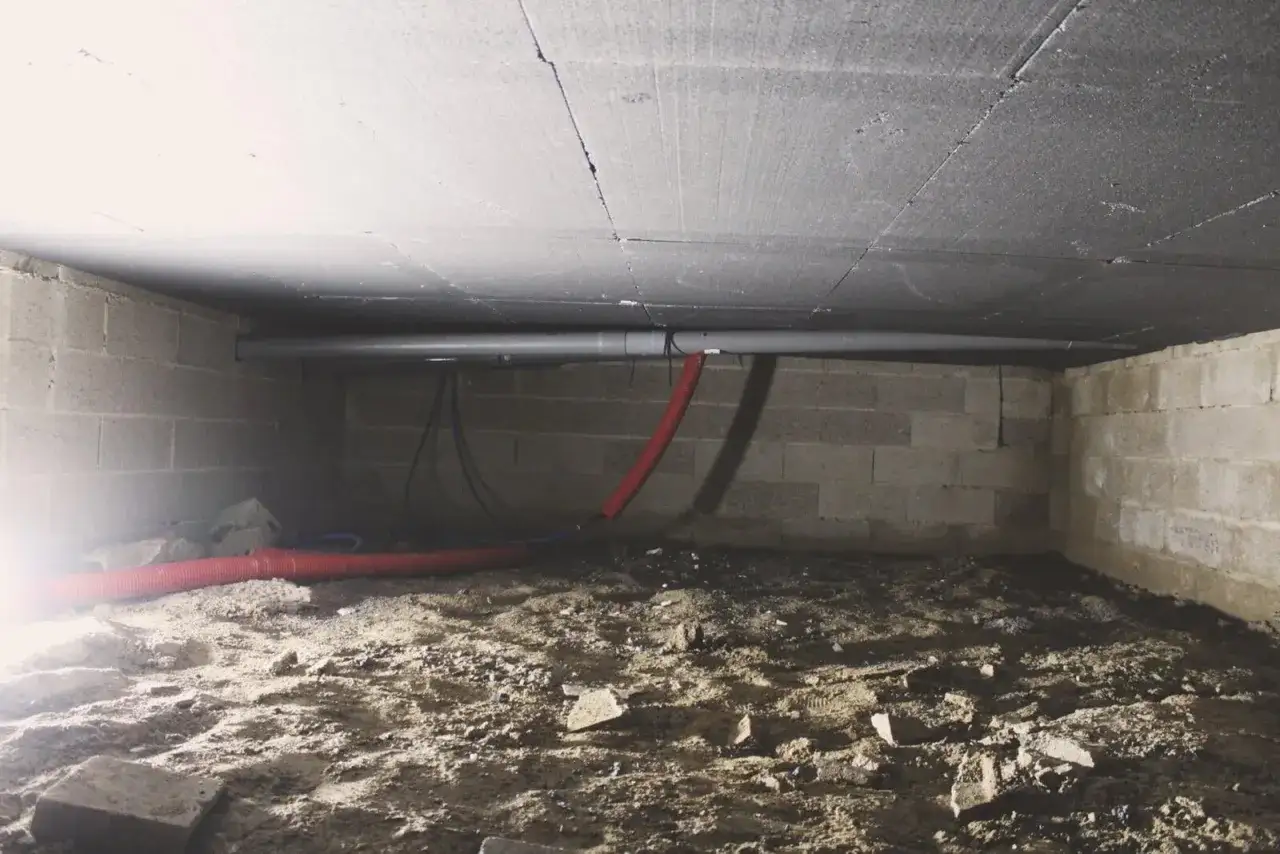 Sous un plafond isolé, des tuyaux traversent un vide sanitaire aux murs de parpaings, le sol est en terre battue.