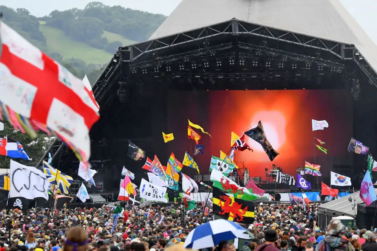 A massive crowd at Glastonbury Festival, with flags waving and a band performing on the main stage. The Glastonbury set times are a hot topic as fans anticipate the performances.