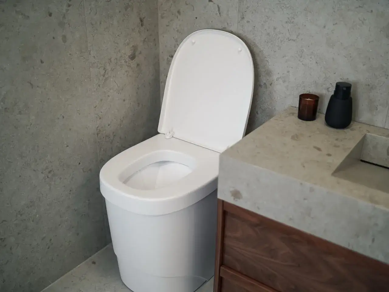 Modern badrum med vit toalett utan avlopp, bredvid ett handfat i betong och tr&auml;.