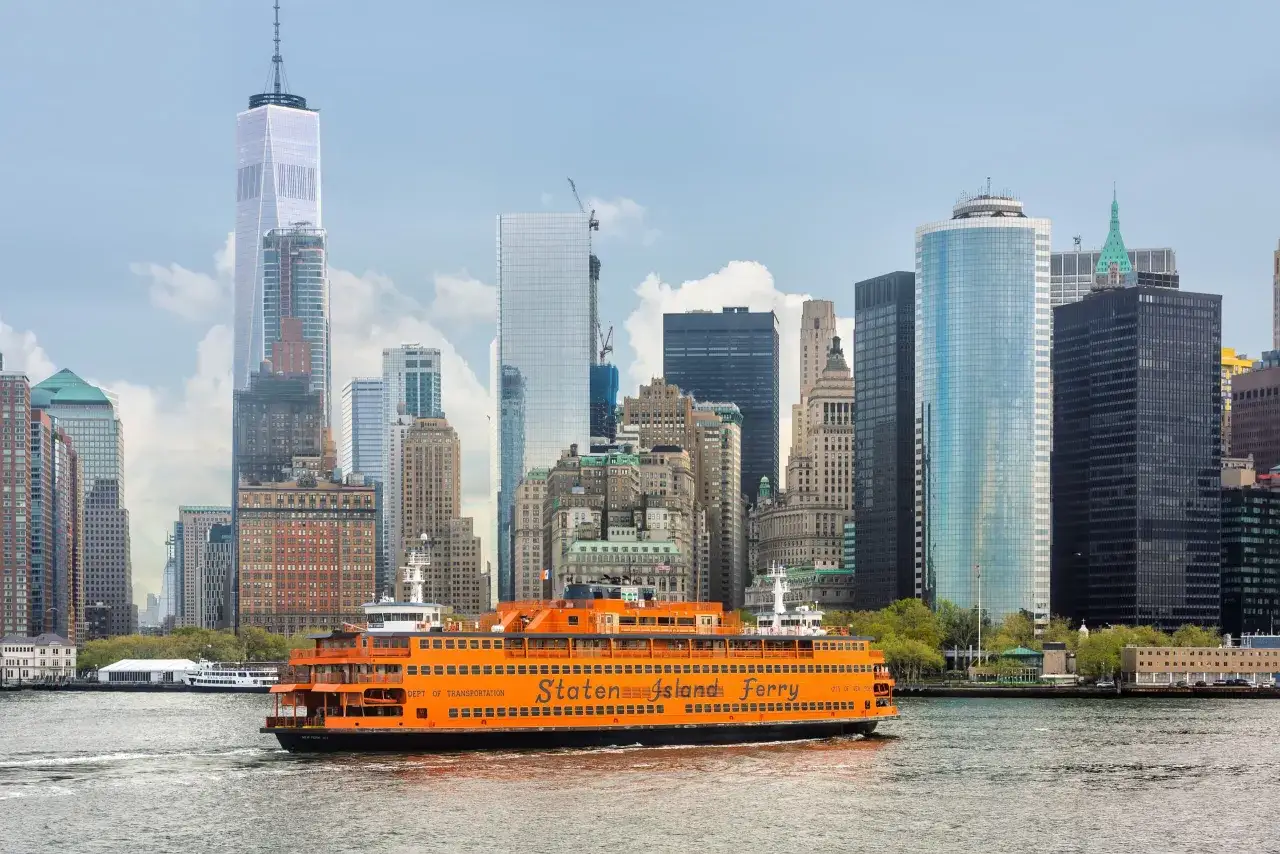 Pomarańczowy prom Staten Island płynie po wodzie na tle drapaczy chmur Manhattanu.