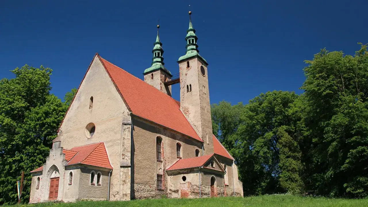 Stary kości&oacute;ł w stolcu, z dwiema wieżami połączonymi mostkiem, otoczony zielenią drzew pod błękitnym niebem.