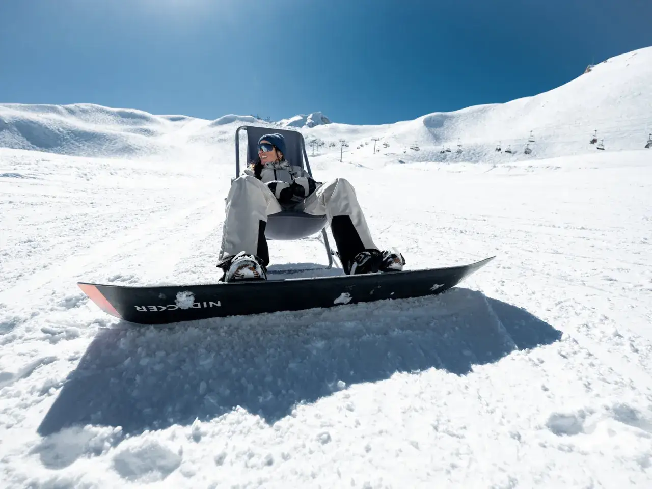Snowboarder détendu sur la neige, se demandant s'il faut farter des skis neufs. Le soleil brille sur les pistes.