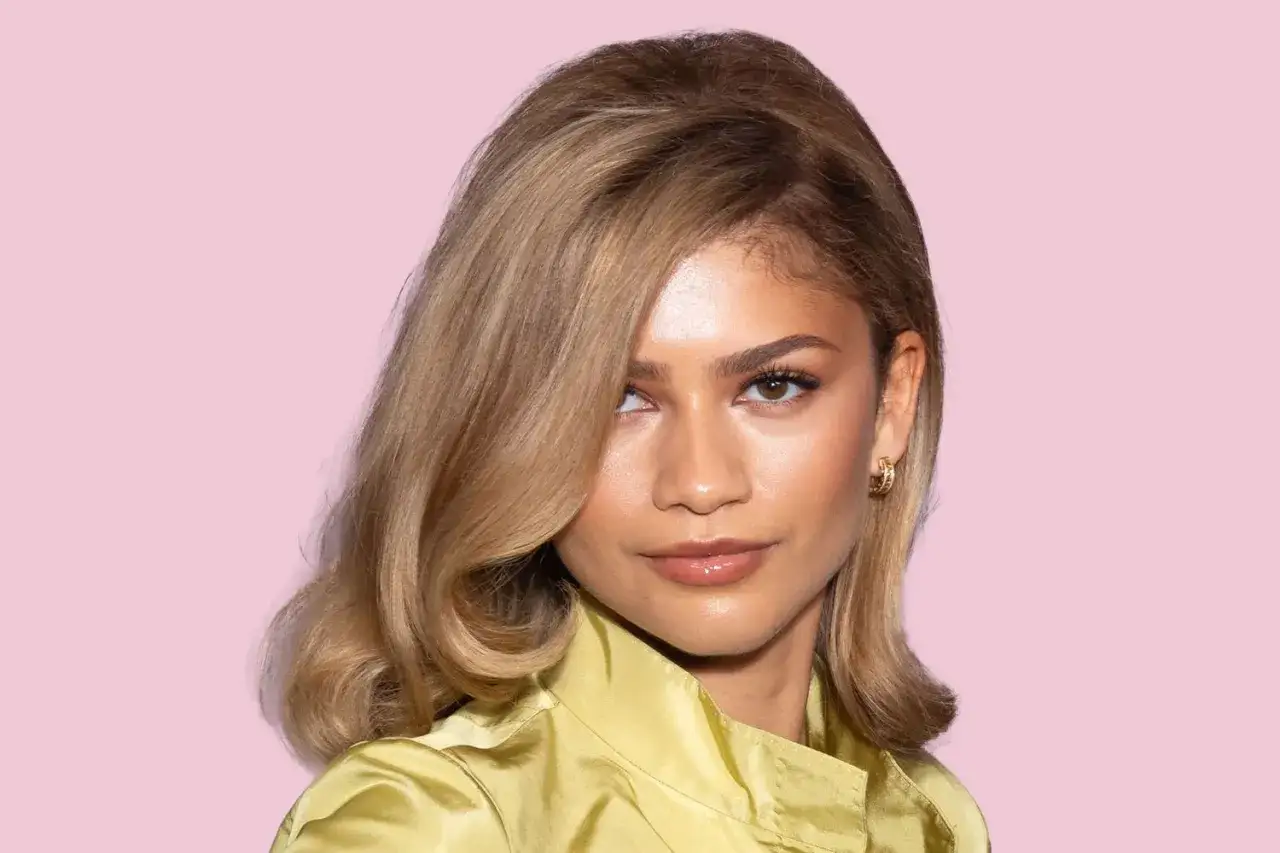 Mujer con cabello rubio sucio, peinado en ondas suaves, con un top amarillo satinado y un fondo rosa.