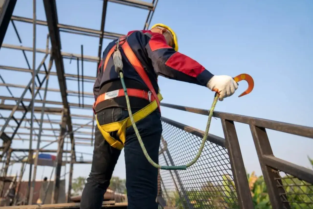 Trabajador con arnés de seguridad y casco amarillo, asegurando su línea de vida. La protección colectiva es esencial en obras.