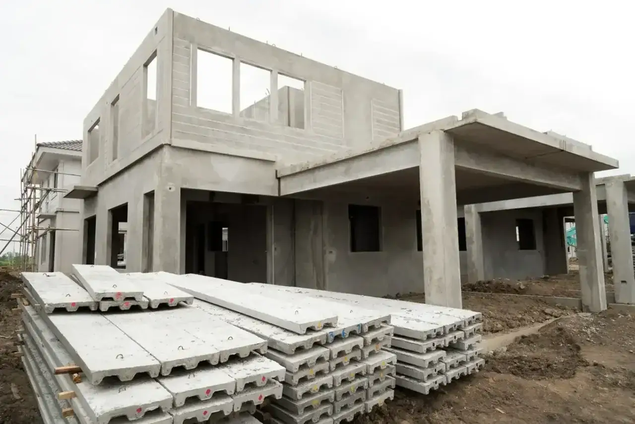 Budowa domu z prefabrykatów betonowych w toku. Na pierwszym planie stosy płyt betonowych, w tle szary, niedokończony budynek.