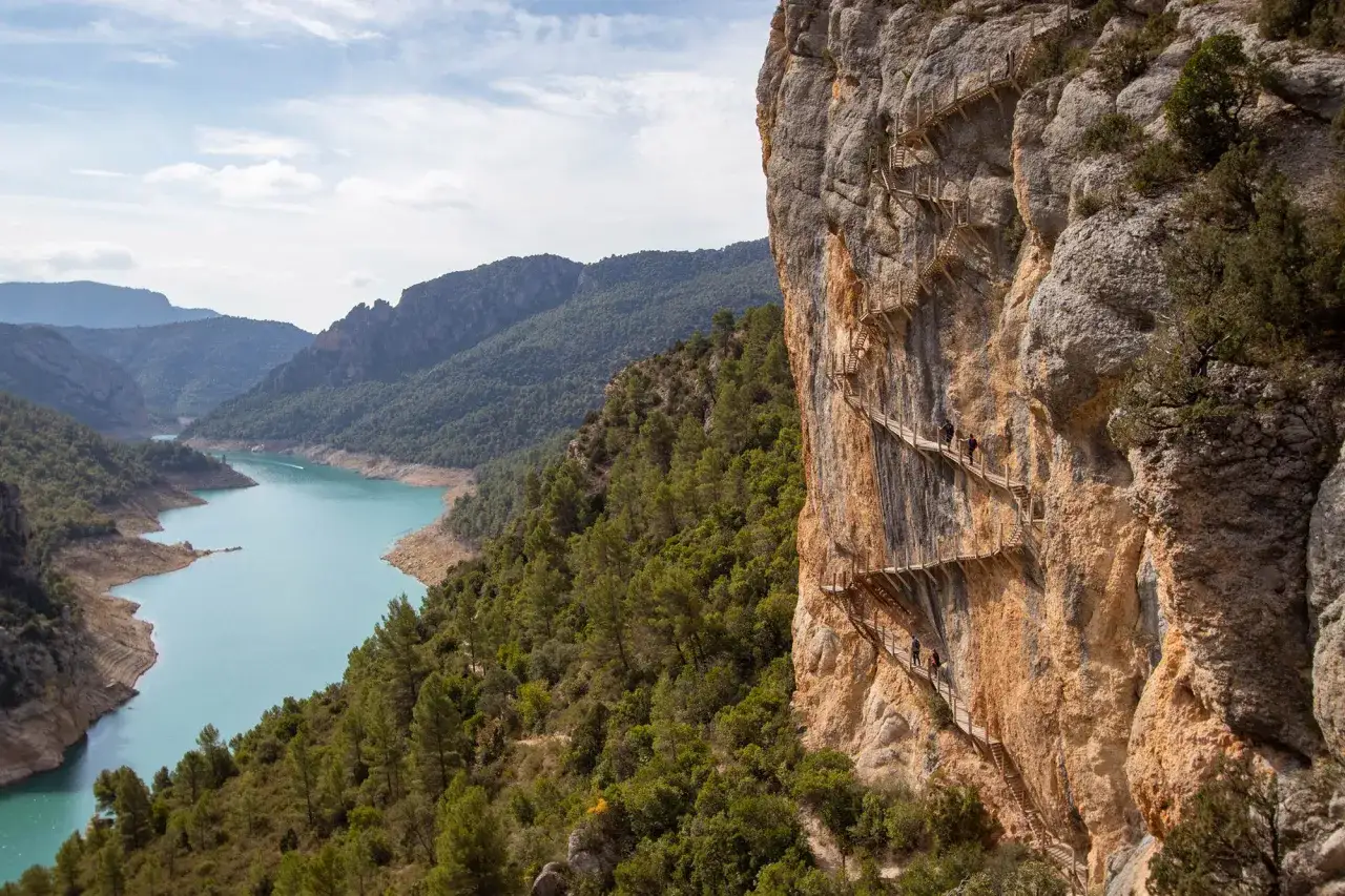 Senderistas exploran el congost de Mont-rebei, un desfiladero con pasarelas de madera en un acantilado sobre un r&iacute;o turquesa.
