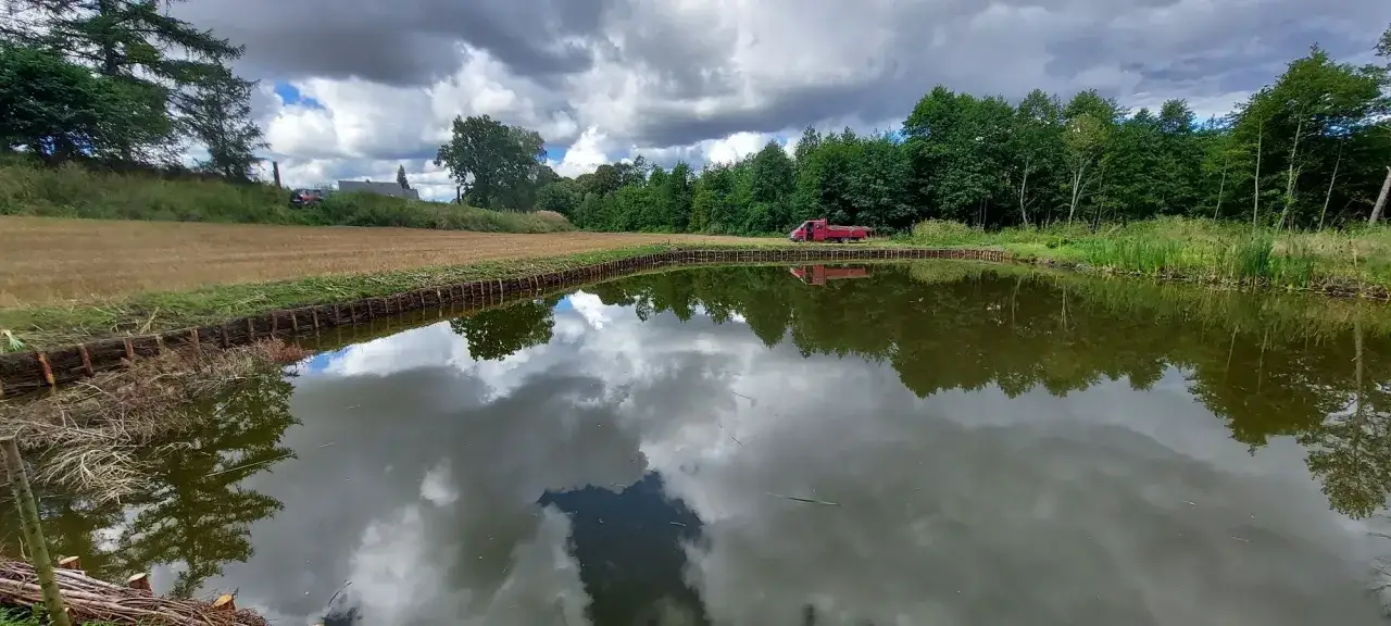 Zbiornik wodny z umocnionym brzegiem za pomocą faszyny, obok pole i las. Czerwony samochód stoi na skarpie.