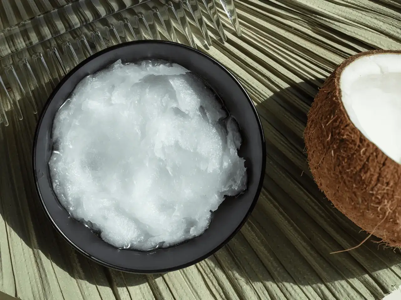 En skål med krämig kokosolja för huden bredvid ett halvt kokosnöt på en palmblad.