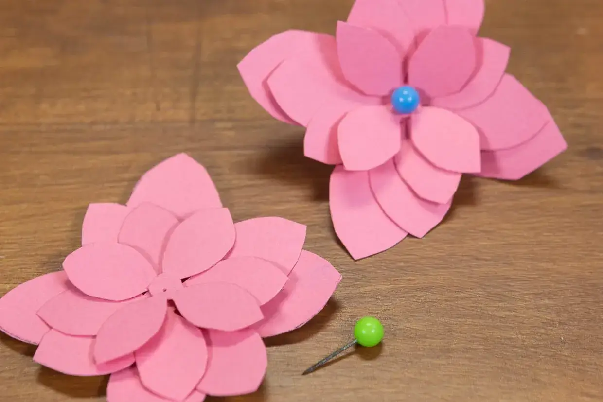 Deux fleurs roses en papier Canson, faciles &agrave; r&eacute;aliser, sur fond de bois. Une petite punaise verte &agrave; c&ocirc;t&eacute;.