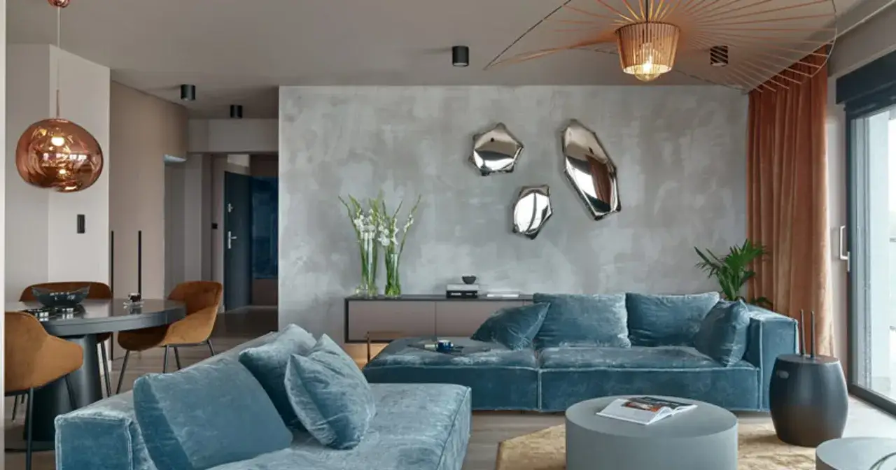 Nowoczesny salon z sofą w odcieniu **kolor lazurowy**. W tle betonowa ściana z ozdobnymi lustrami i designerska lampa.