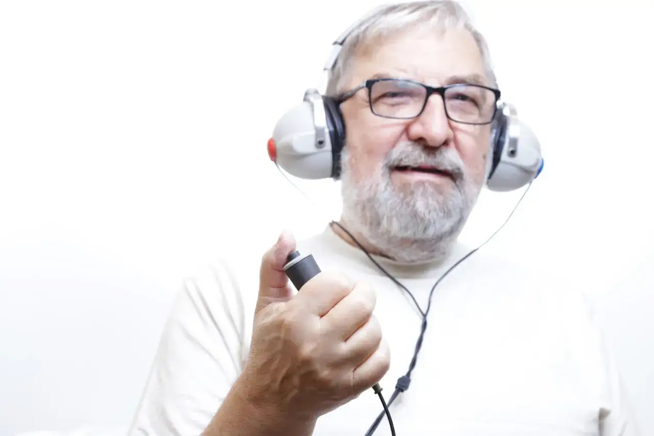 Starszy mężczyzna w okularach i słuchawkach trzyma przycisk, być może podczas badania słuchu. Ubytek słuchu 30 dB.