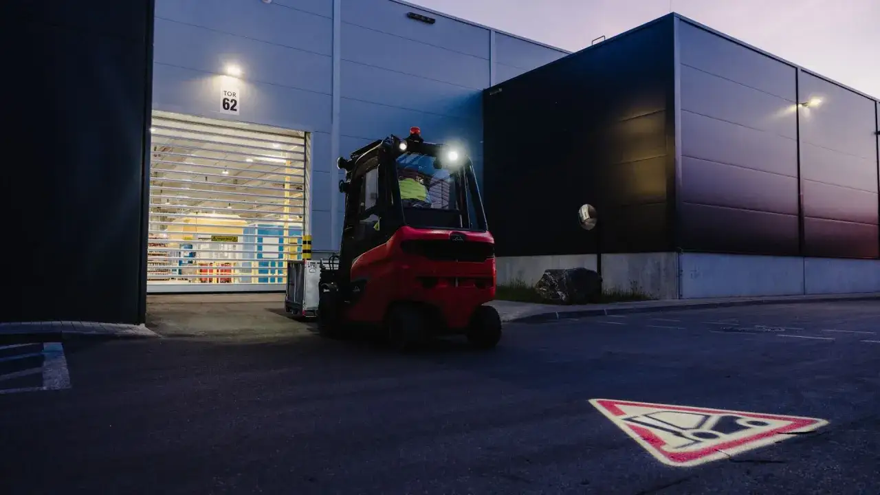 En röd truck med stark belysning står vid en lagerport. Det är mörkt ute, vilket visar hur viktigt det är att en truck har belysning för säkerheten.