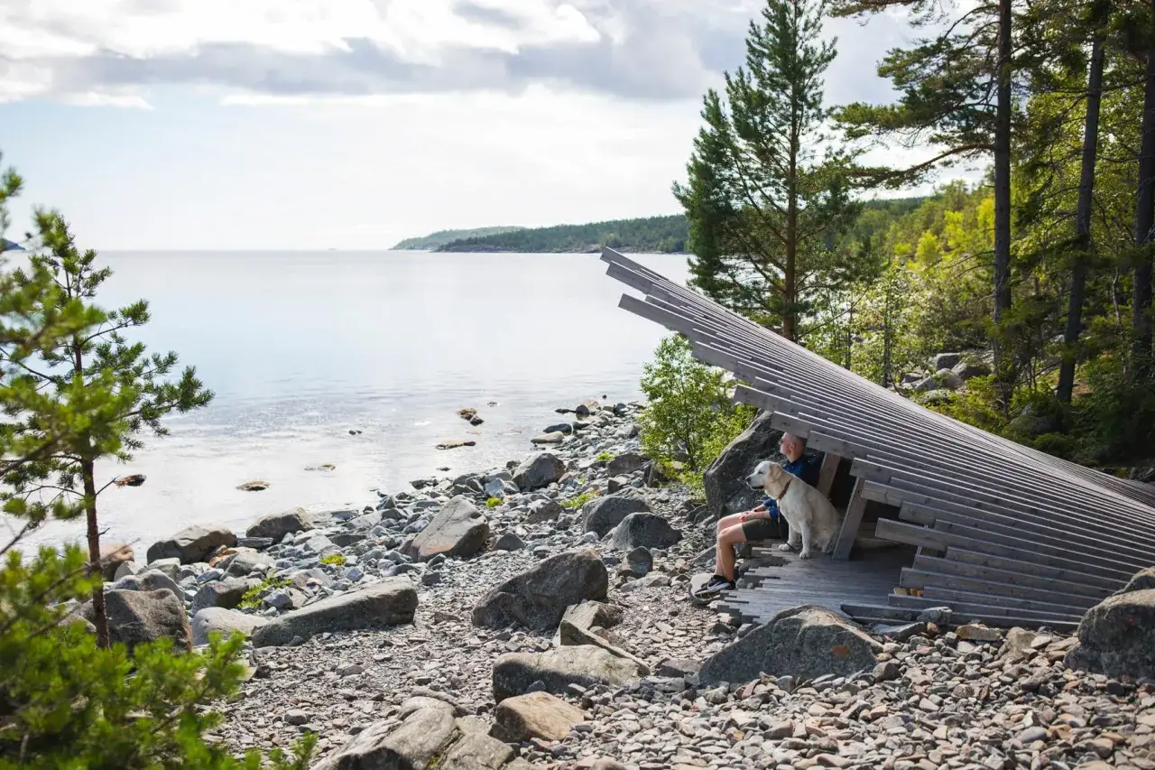 Man och hund njuter av en lugn semester vid havet, sittandes under ett unikt tr&auml;konstruktion p&aring; en stenig strand.