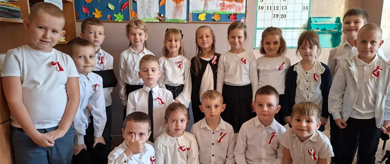 Dzieci w białych koszulach i bluzkach z biało-czerwonymi wstążeczkami, gotowe do wypełniania obowiązku szkolnego.