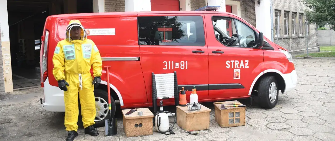 Strażak w żółtym kombinezonie gotowy do akcji. Obok czerwony wóz strażacki i sprzęt, który może być użyty, gdy straż pożarna usuwa gniazda os.
