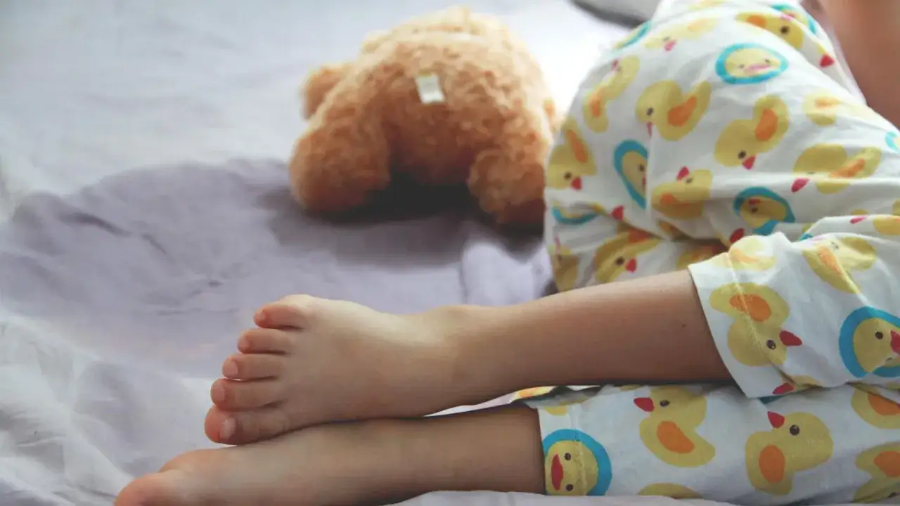 F&uuml;&szlig;e eines Kindes in Enten-Pyjama auf einem Bett. Ein Teddyb&auml;r liegt daneben. Hoffentlich bleibt das Kind nachts trocken.