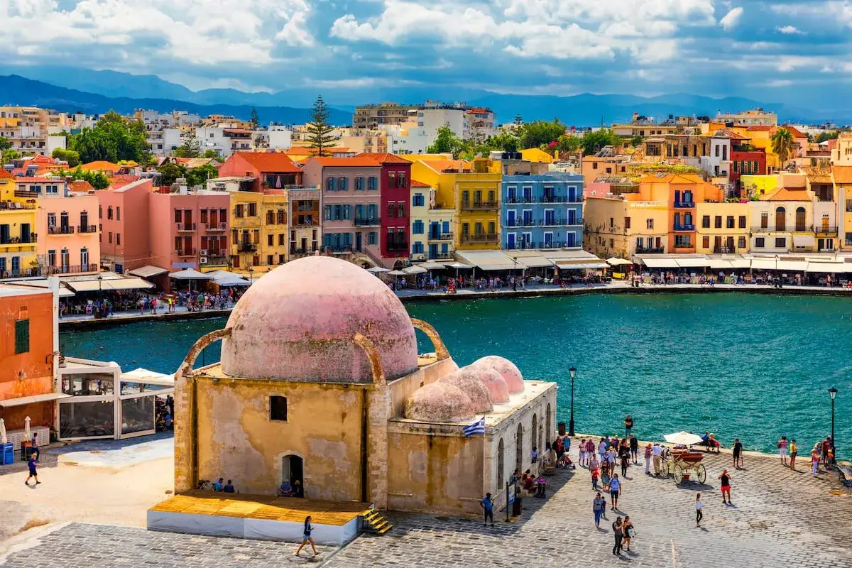 Puerto de Chania, Creta que ver: edificios coloridos, un domo rosado y el mar azul. Gente paseando por el muelle.