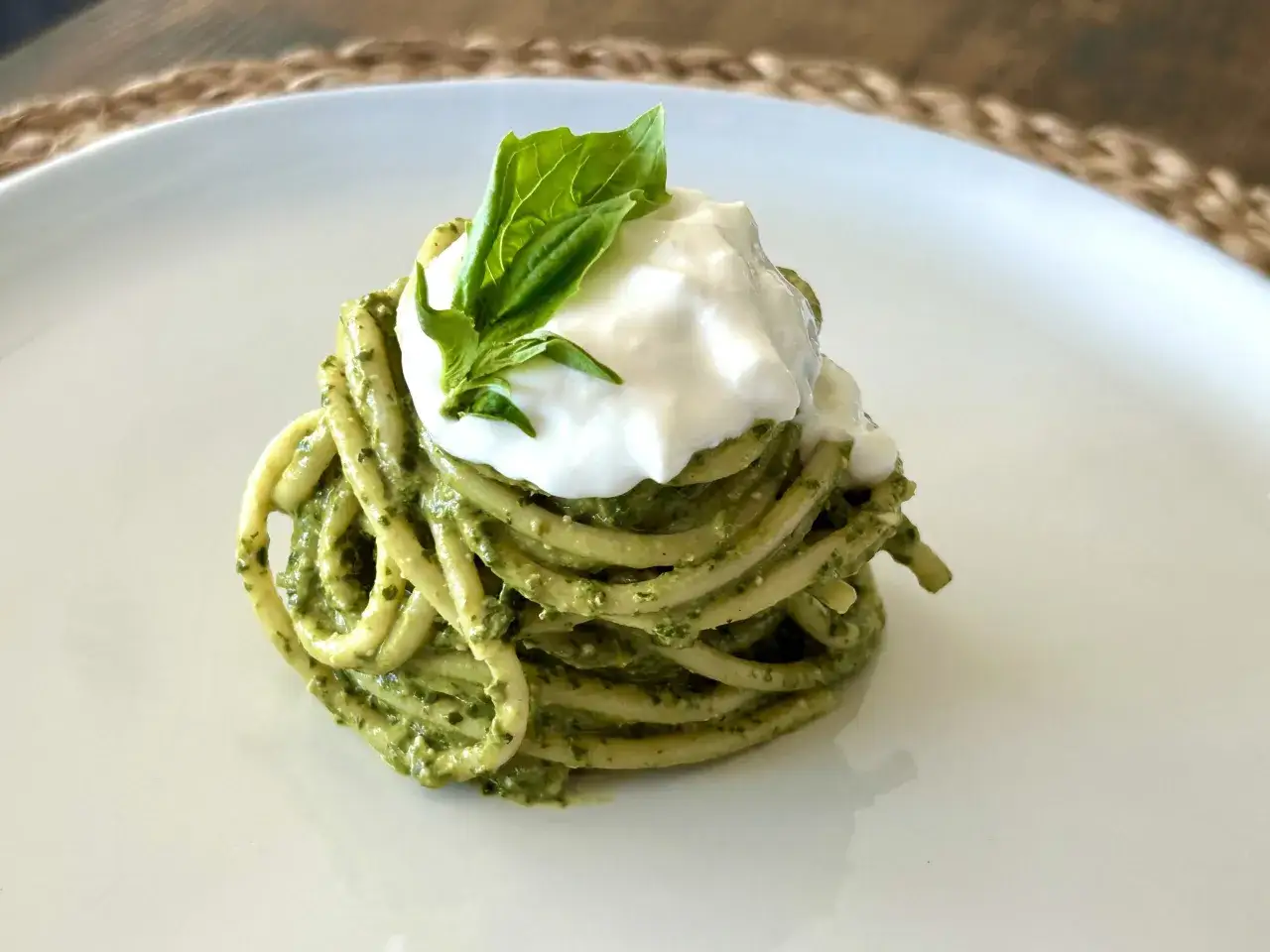 Pâtes spaghetti enroulées, nappées d'une sauce pesto verte vibrante, surmontées d'une boule de mozzarella crémeuse et d'une feuille de basilic frais.