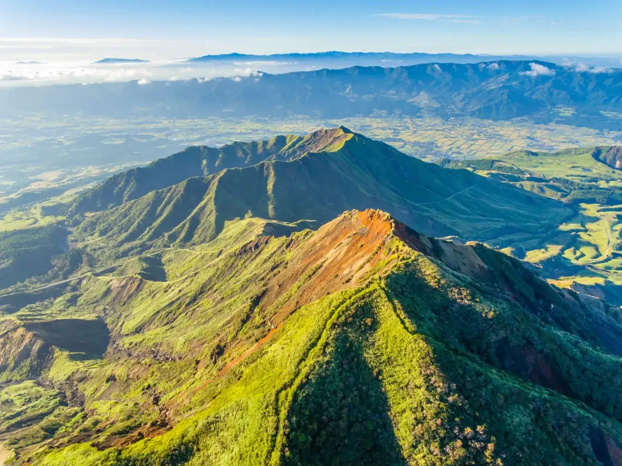 Vue aérienne d'un **aso volcan** verdoyant, ses crêtes déchiquetées baignées de soleil, dominant une vallée fertile et des montagnes lointaines.