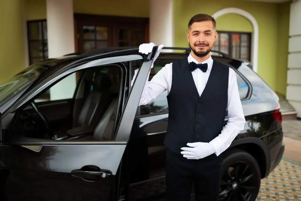 Uśmiechnięty kamerdyner w białych rękawiczkach otwiera drzwi luksusowego czarnego samochodu, oferując usługi **valet parking**.