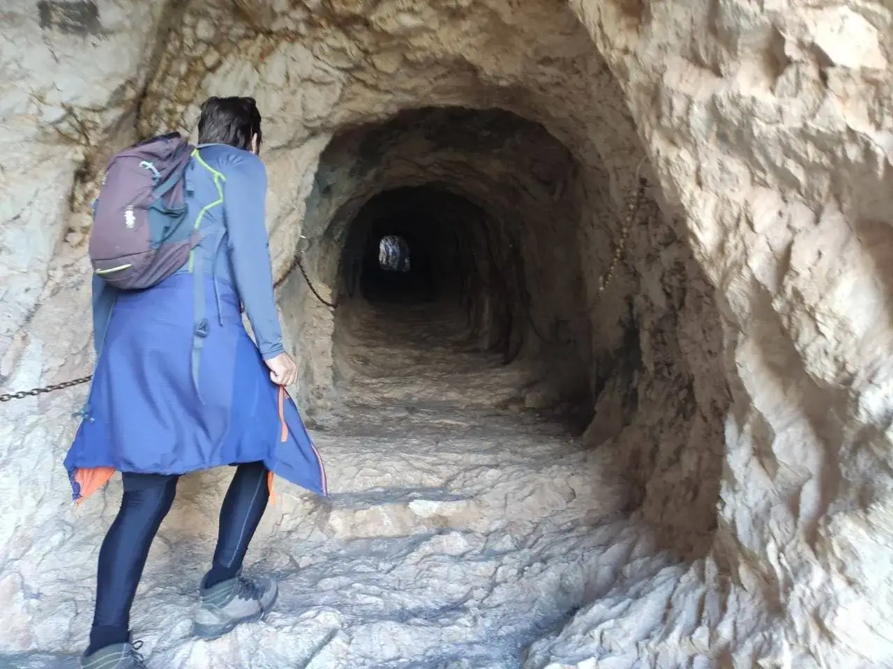 Persona camina por un t&uacute;nel rocoso en la ruta del Pe&ntilde;&oacute;n de Ifach, con cadenas a los lados.