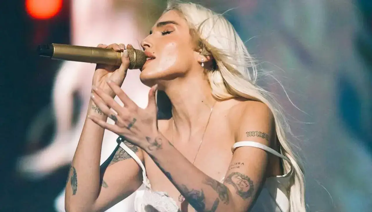 Elena Rose, with her blonde hair flowing, sings passionately into a golden microphone, her tattooed arms gesturing expressively.