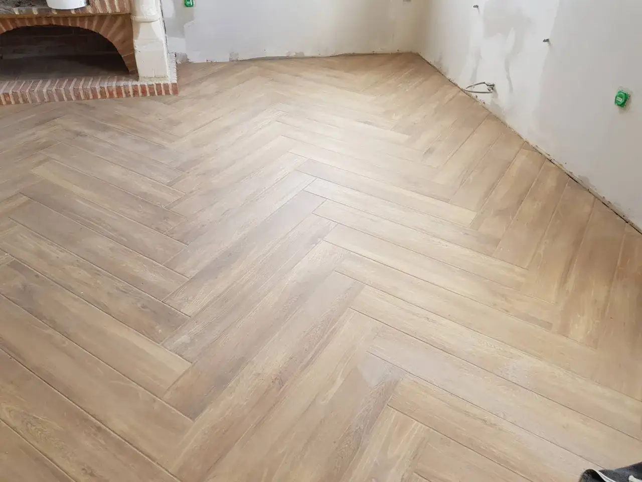 Un sol en bois clair, pos&eacute; en chevron, apporte une touche chaleureuse &agrave; cette pi&egrave;ce en cours d'am&eacute;nagement, pr&egrave;s d'une chemin&eacute;e en briques.