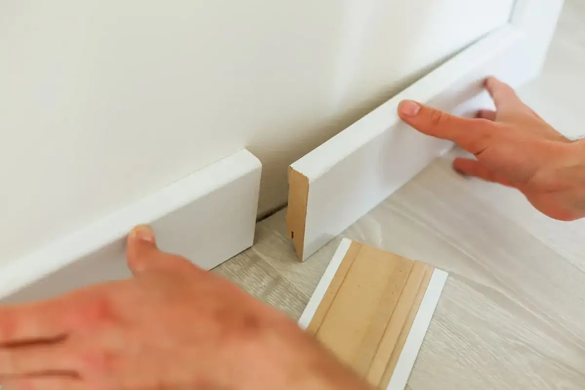 H&auml;nde kleben wei&szlig;e Sockelleisten an die Wand. Ein St&uuml;ck Leiste liegt bereit.