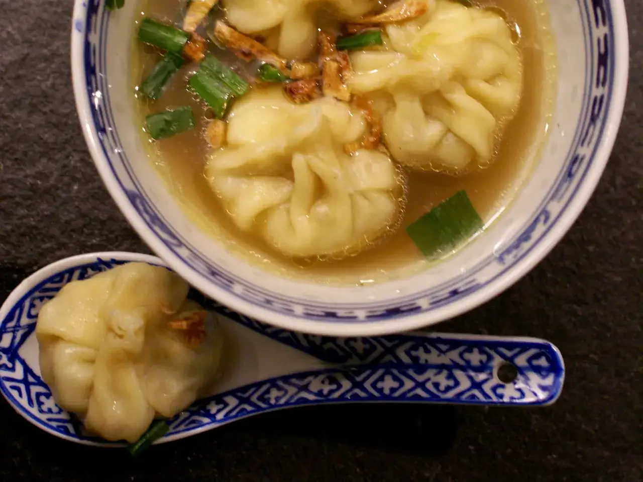 A miseczce z niebiesko-białym wzorem pływa aromatyczna zupa wonton z zieloną cebulką i chrupiącymi dodatkami. Na łyżce leży jeden pierożek wonton.