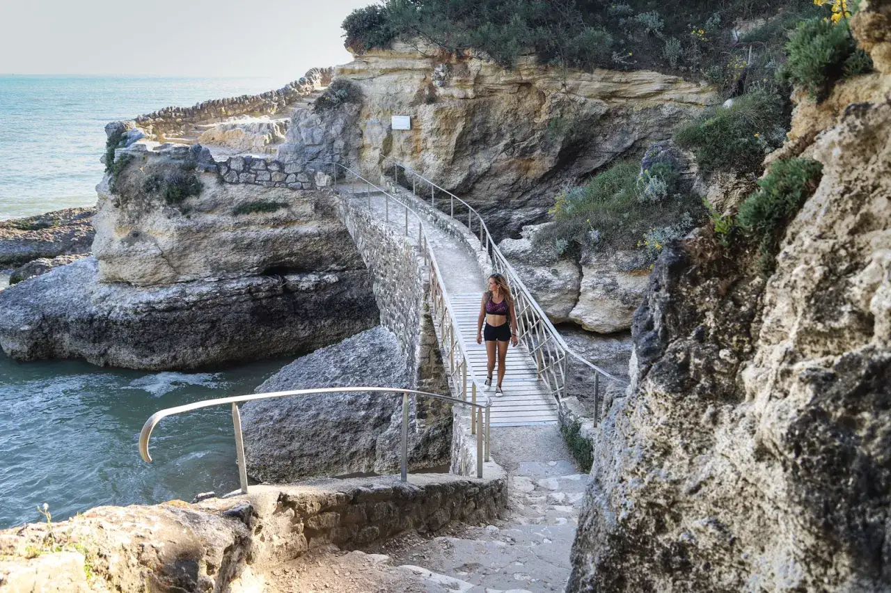 Une femme profite d'un trek en Bretagne, traversant un pont en pierre au-dessus de l'eau turquoise, entourée de falaises rocheuses.