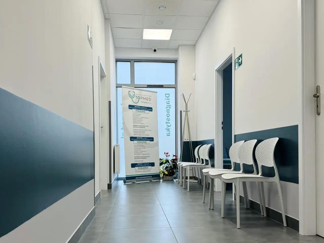 Nowoczesny korytarz przychodni medycznej z białymi krzesłami i banerem informacyjnym.