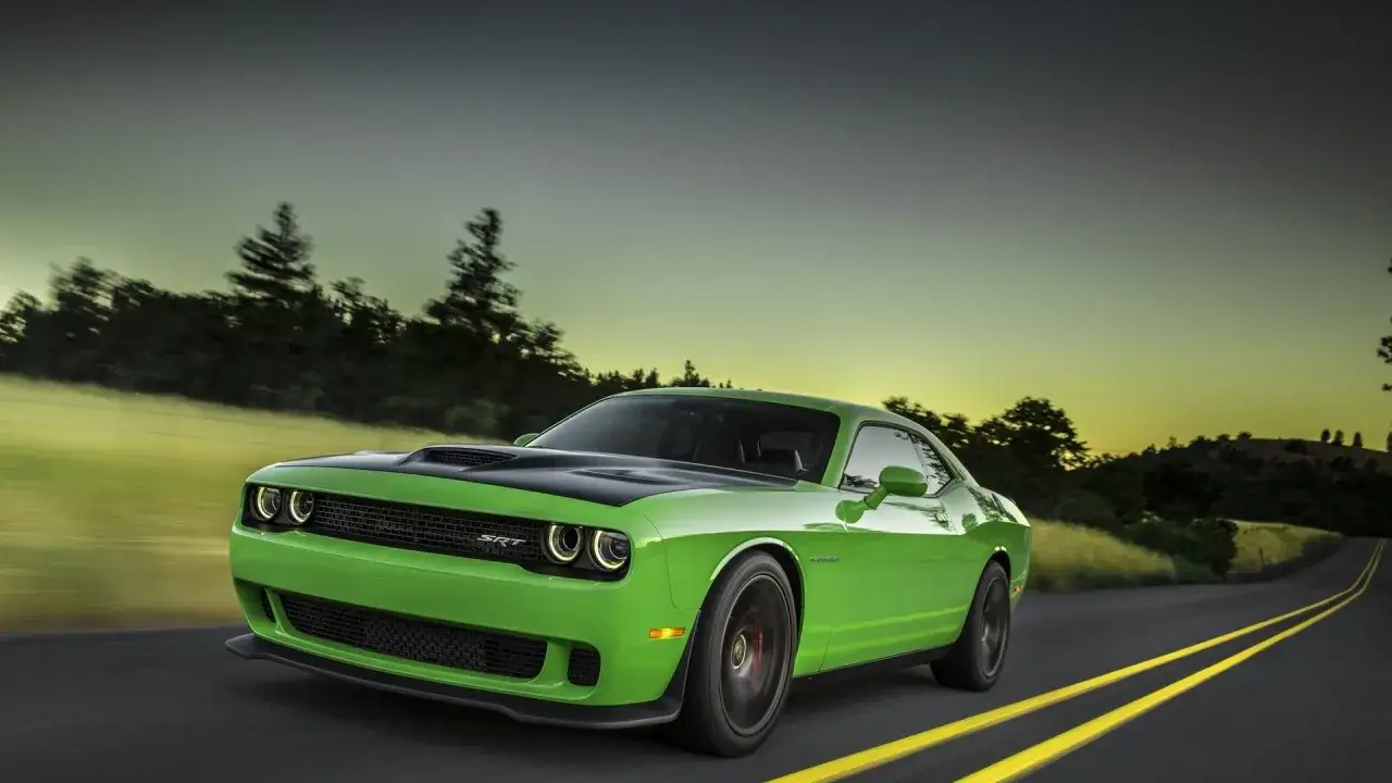 Jaskrawozielony Dodge Challenger Hellcat pędzi po pustej drodze o zachodzie słońca, otoczony drzewami i wzgórzami.