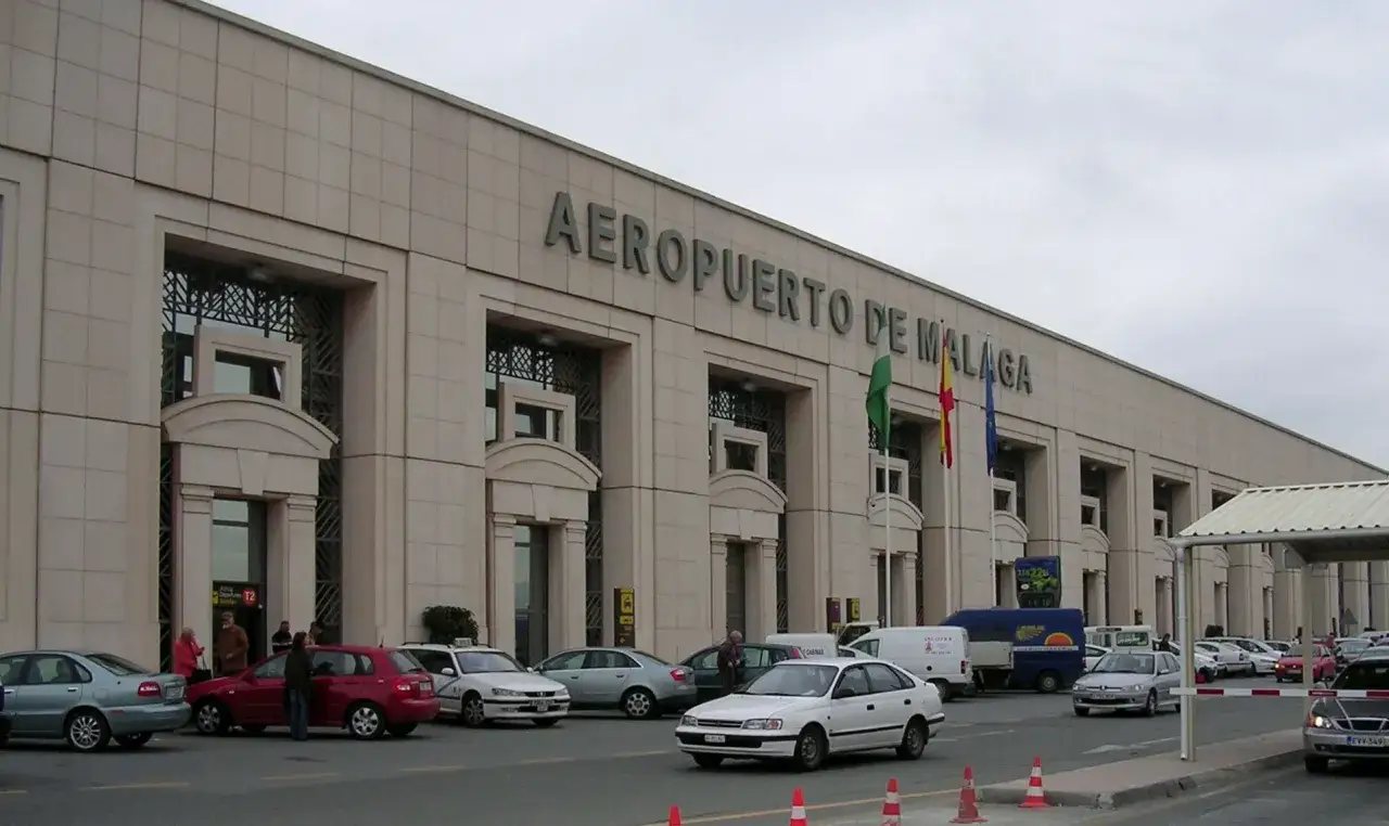 Budynek Aeropuerto de Malaga, z którego odlatują samoloty. Przed budynkiem parkują samochody, a obok powiewają flagi.