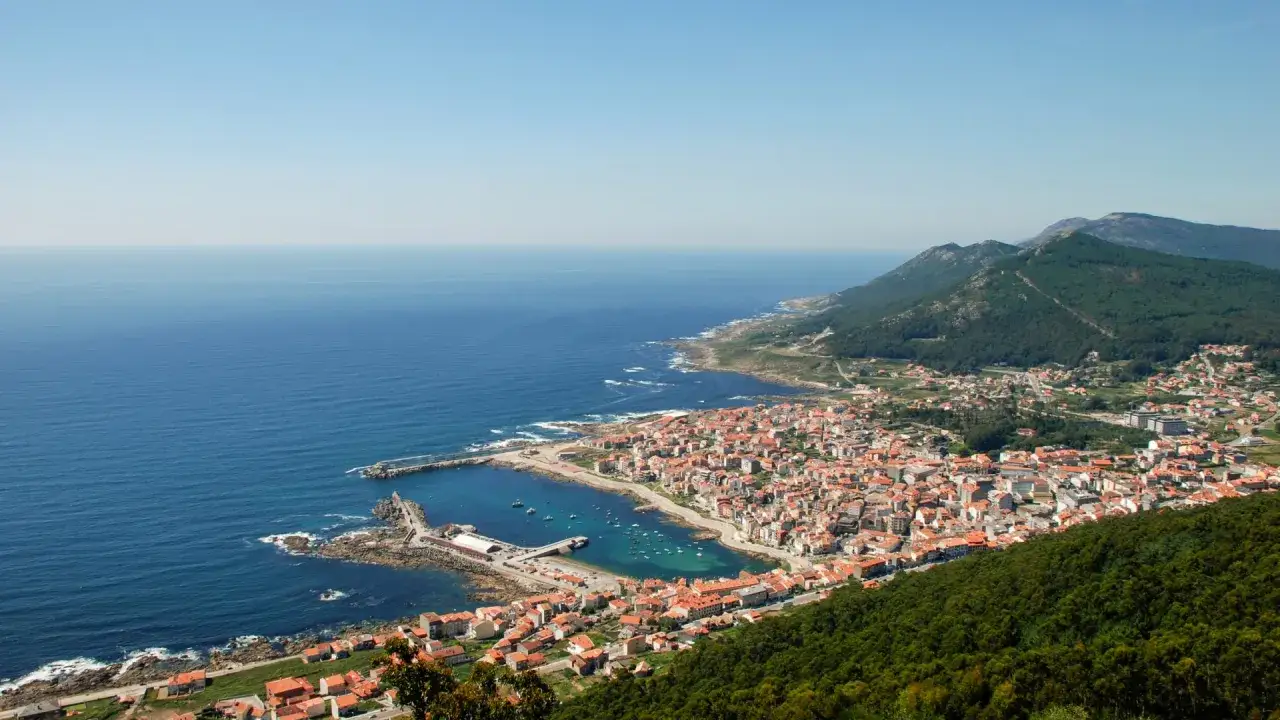Vista aérea de un pueblo costero con un puerto lleno de barcos, ideal para la pesca. El mar azul se extiende hasta el horizonte.