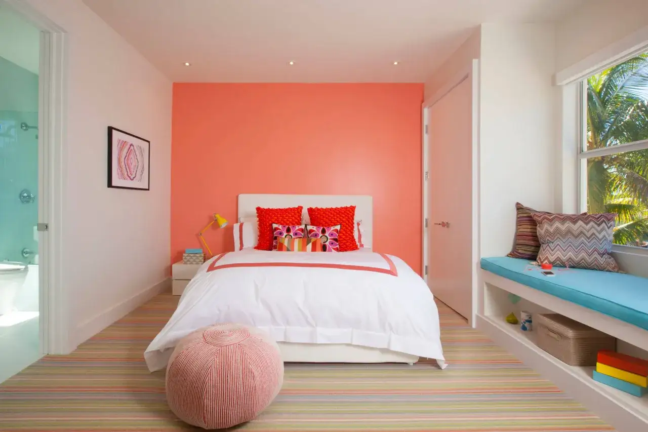 Chambre lumineuse avec mur couleur corail, lit blanc, tapis ray&eacute; et pouf. Un banc pr&egrave;s de la fen&ecirc;tre offre une vue sur la v&eacute;g&eacute;tation tropicale.