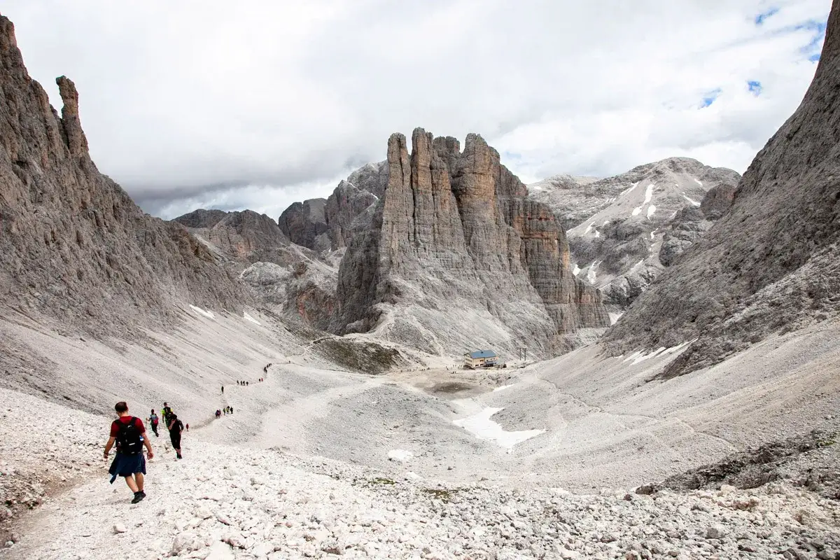 Escursionisti si dirigono verso le imponenti Torri del Vajolet, un paesaggio roccioso e maestoso sotto un cielo nuvoloso.