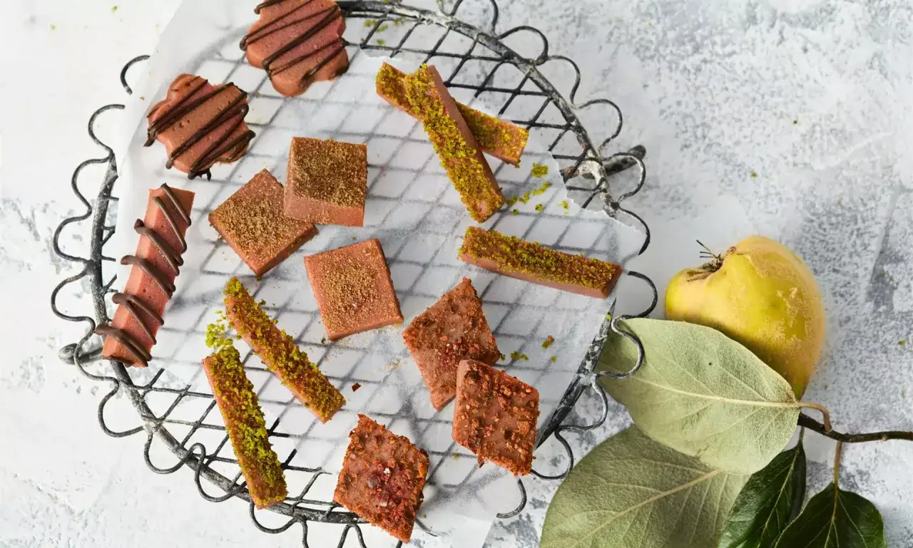 Leckeres Quittenbrot-Rezept: Rosa Quadrate und Stäbchen mit Pistazien und Schokolade auf einem Gitter. Daneben eine Quitte.