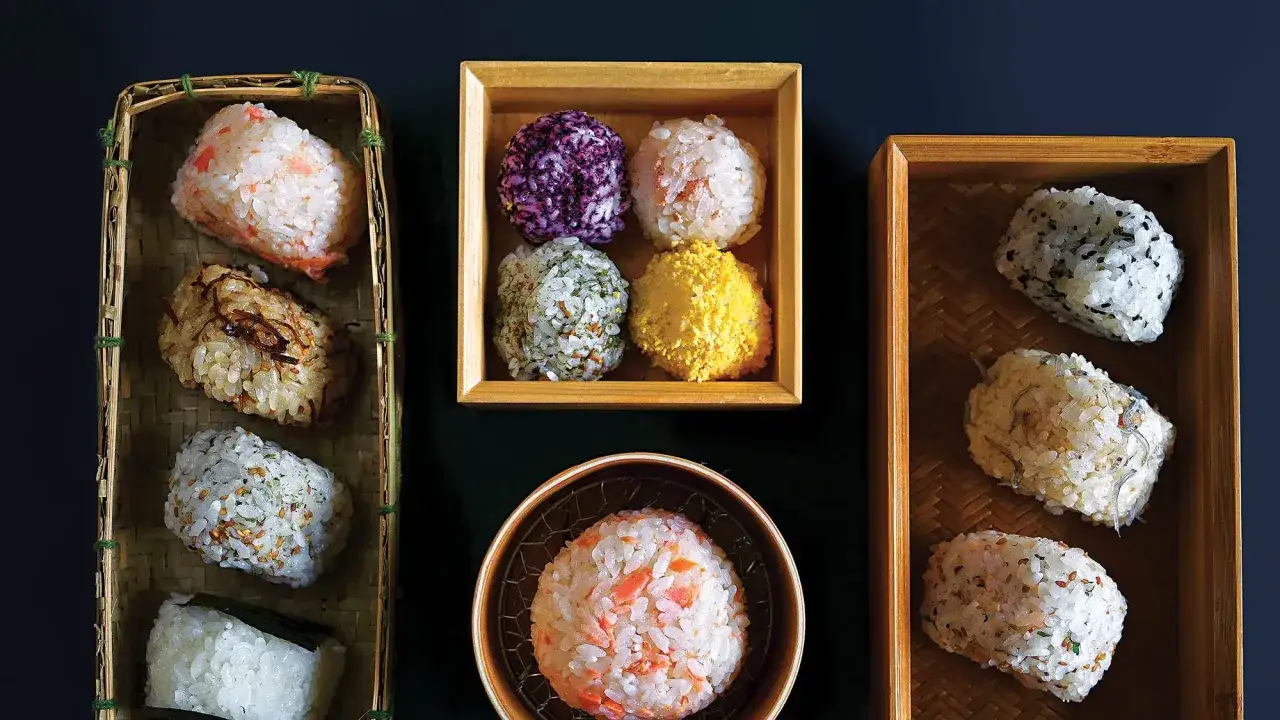 Różnorodne japońskie przekąski z ryżu, inspirowane przepisami na onigiri. W koszykach i pudełkach widać kulki ryżowe z dodatkami: łososiem, sezamem, ziołami, a nawet fioletowym ryżem.