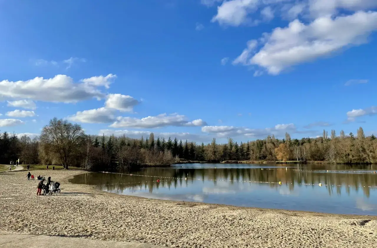 Plage de sable fin au bord d'un lac paisible, avec des arbres en arrière-plan rappelant la forêt de Fontainebleau. Une famille profite du soleil.