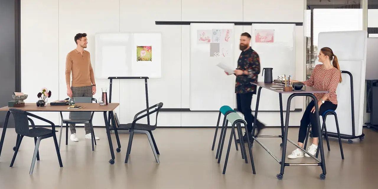 Une &eacute;quipe dynamique participe &agrave; une r&eacute;union debout, &eacute;changeant des id&eacute;es autour de tableaux blancs et de tables hautes.