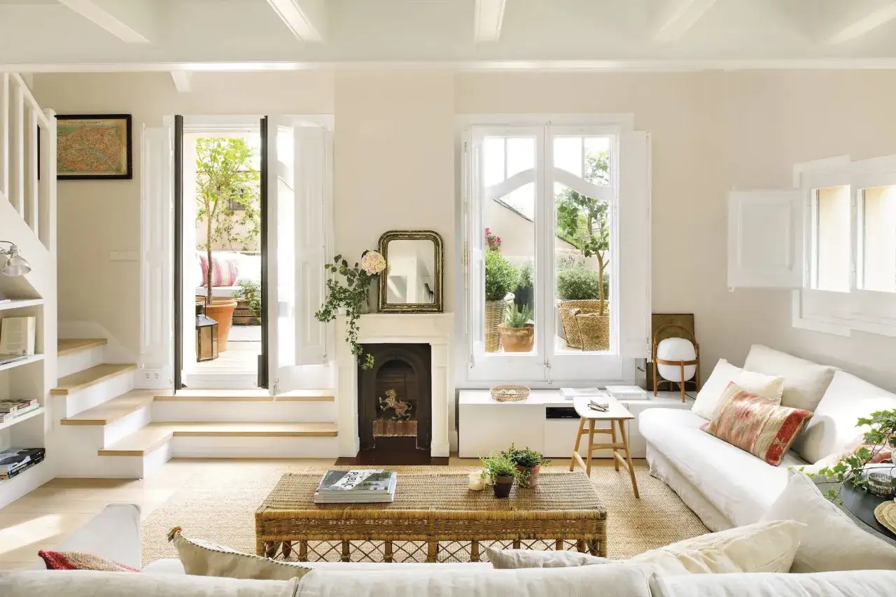Salón luminoso con chimenea, sofá blanco y mesa de mimbre. La pared, pintada en un suave tono ral 9010, realza la calidez del espacio.