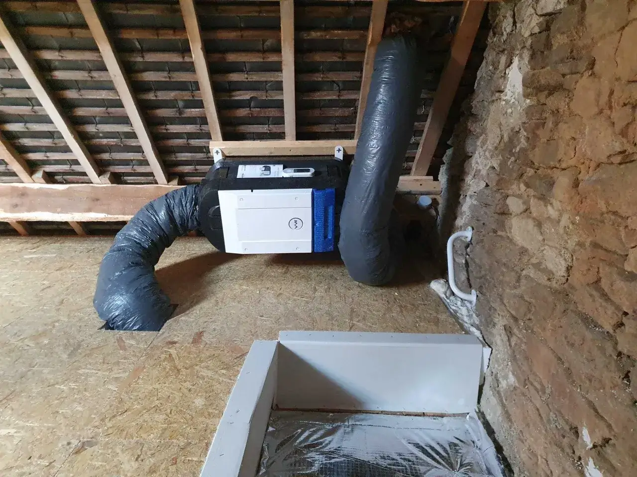 Système de ventilation par surpression installé dans un grenier, avec des conduits souples gris et une unité blanche fixée aux poutres.