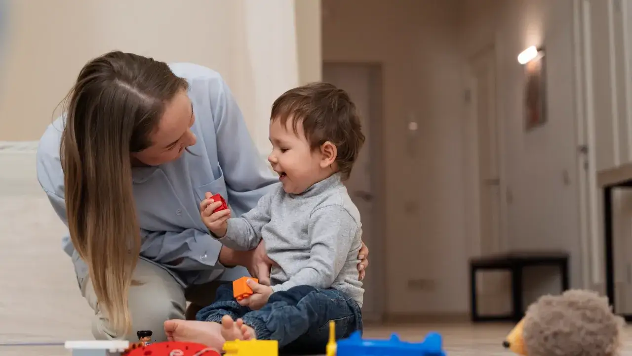 Madre e hijo juegan con bloques. La madre, en un gesto de adaptaci&oacute;n de jornada por cuidado de hijos, se agacha para compartir este momento.