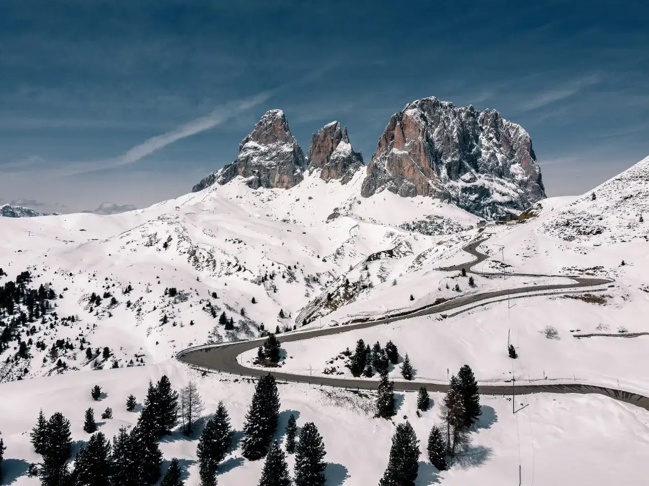 Strada tortuosa tra i **passi delle Dolomiti** innevate, con imponenti vette rocciose sullo sfondo e pini solitari.